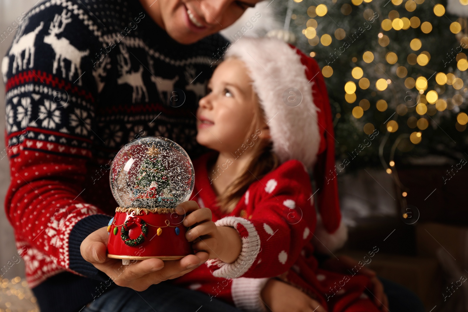 Photo of Father and daughter with snow globe near Christmas tree, focus on toy