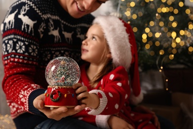 Photo of Father and daughter with snow globe near Christmas tree, focus on toy