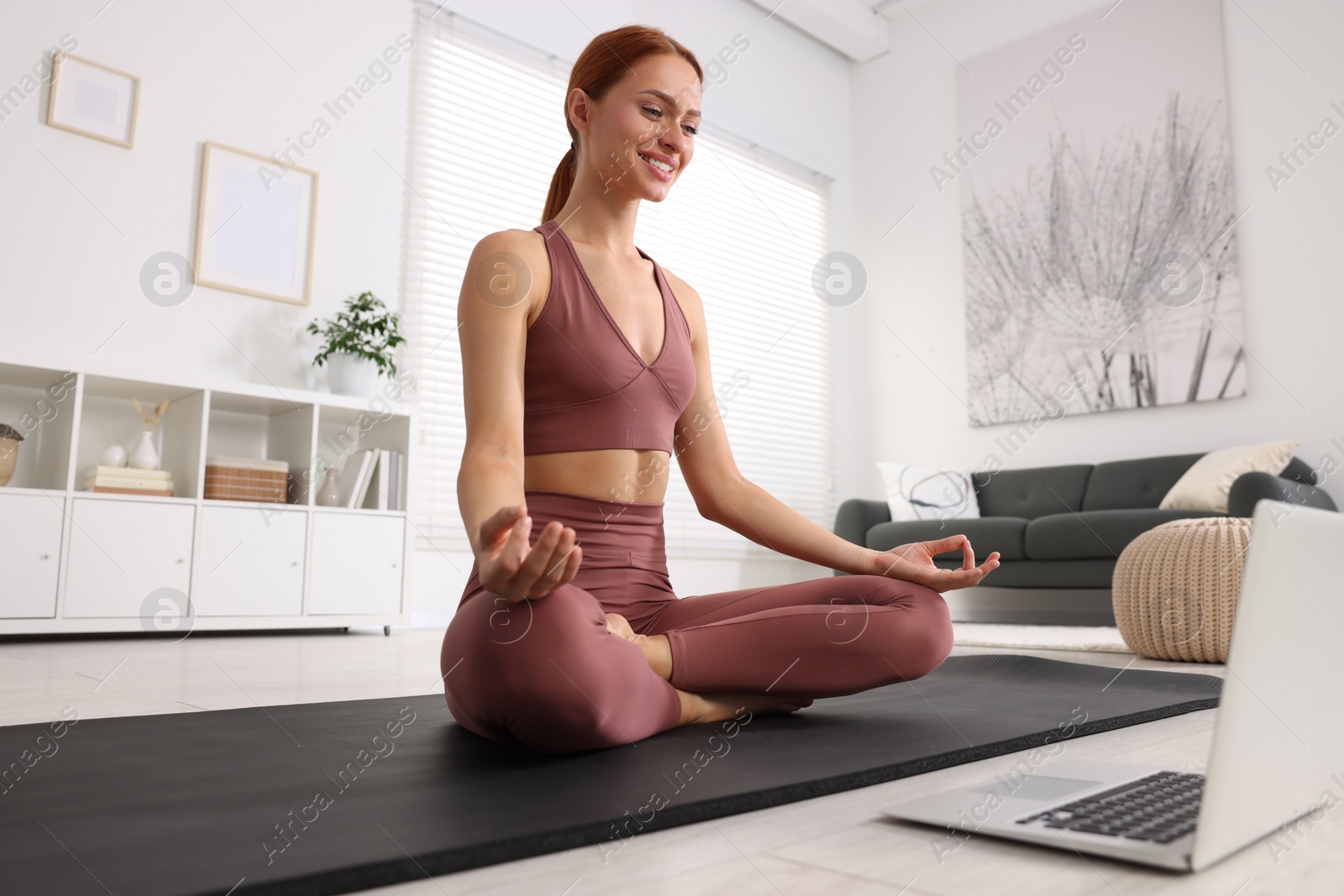 Photo of Beautiful young woman practicing Padmasana while watching online class on yoga mat at home. Lotus pose