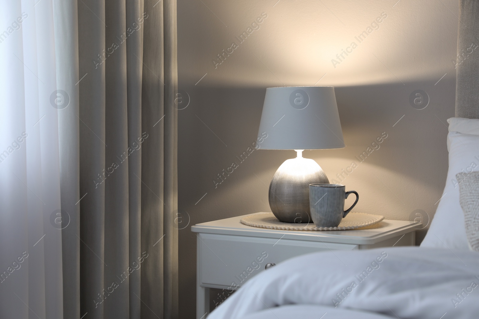 Photo of Stylish lamp and cup of drink on white nightstand in bedroom, space for text