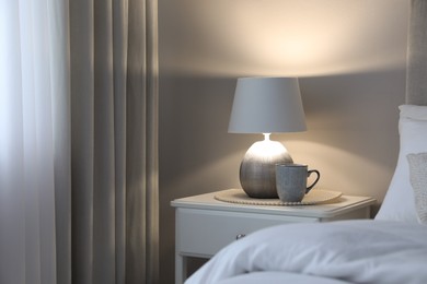 Stylish lamp and cup of drink on white nightstand in bedroom, space for text