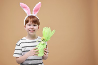 Easter celebration. Cute little boy with bunny ears and wrapped egg on dark beige background. Space for text