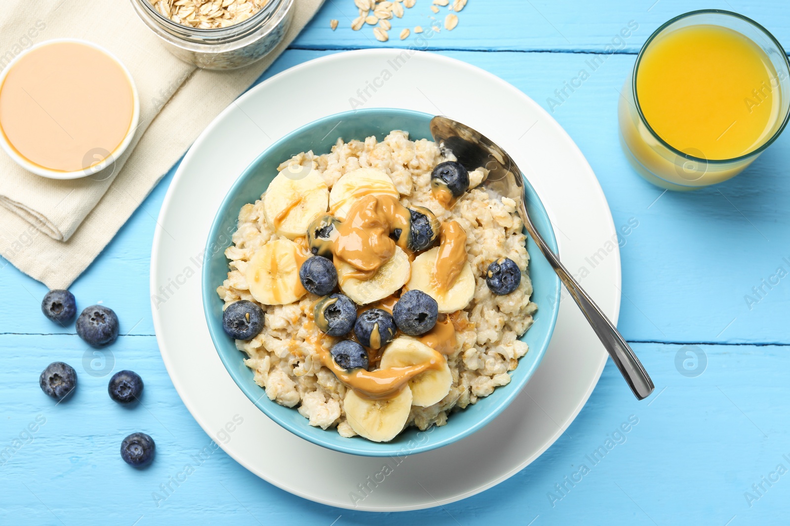 Photo of Tasty oatmeal with banana, blueberries and peanut butter served in bowl on light blue wooden table, flat lay