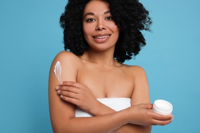 Young woman applying body cream onto shoulder on light blue background