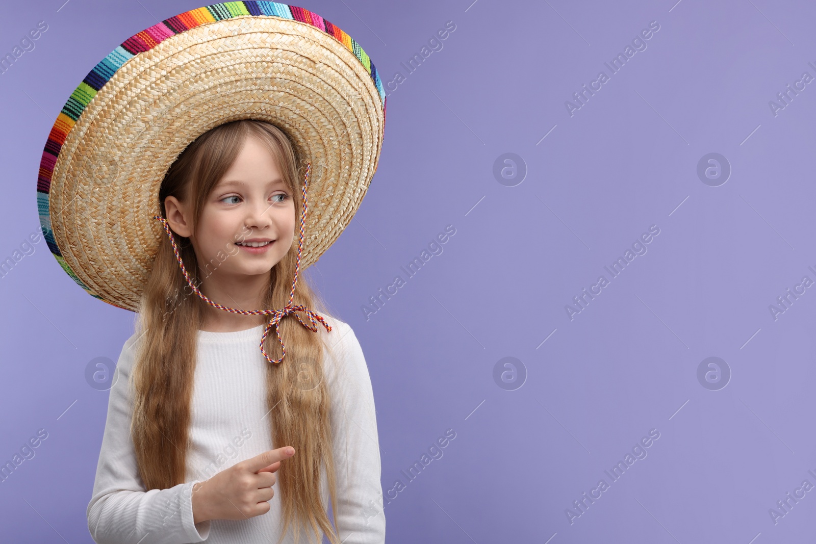 Photo of Cute girl in Mexican sombrero hat pointing at something on purple background. Space for text
