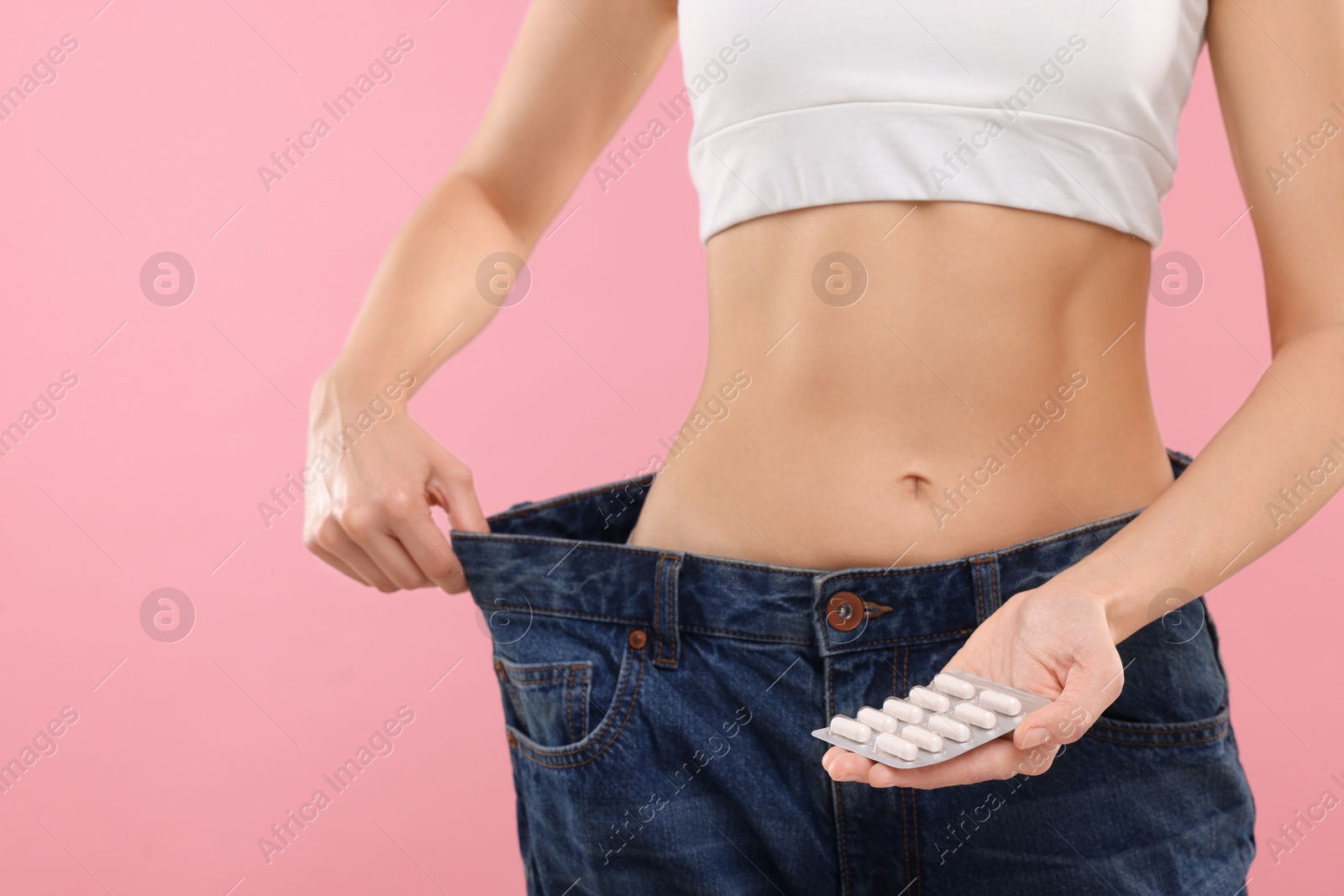 Photo of Slim woman in big jeans with pills on pink background, closeup. Weight loss