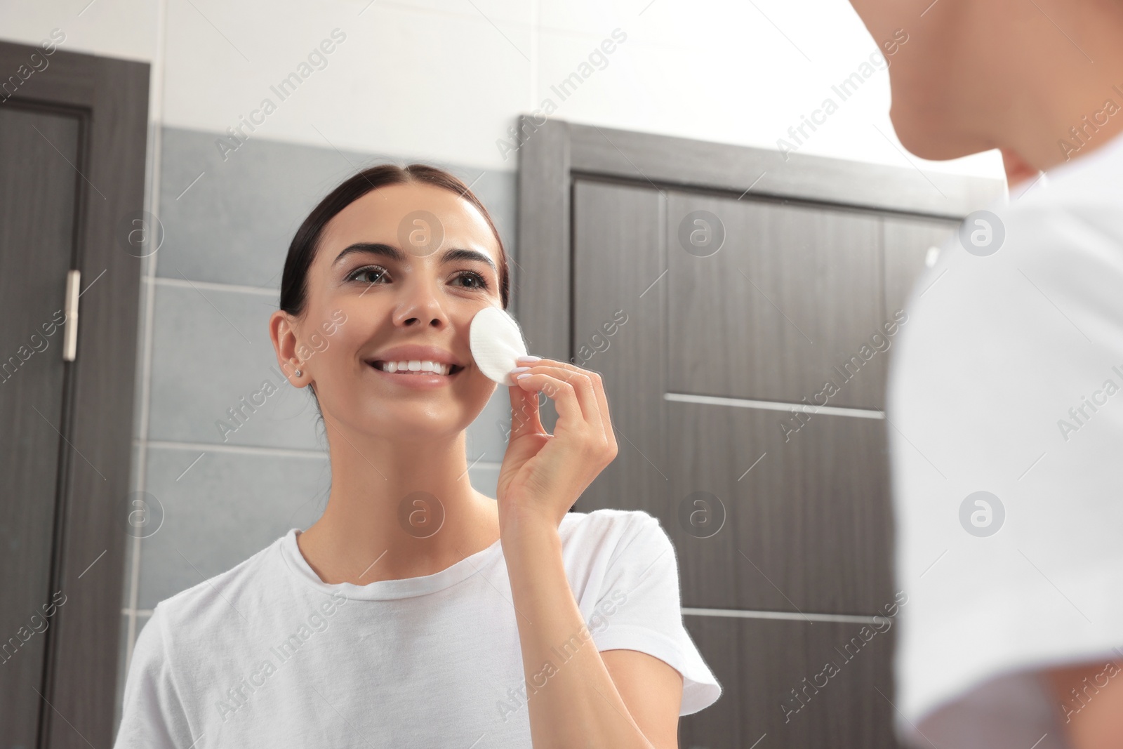 Photo of Young woman using cotton pad with micellar water near mirror in bathroom