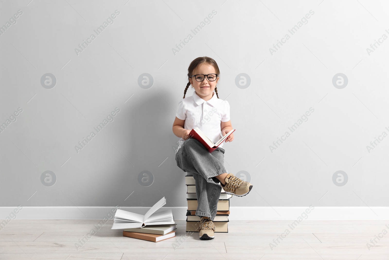 Photo of Cute little girl in glasses sitting on stack of books near light grey wall