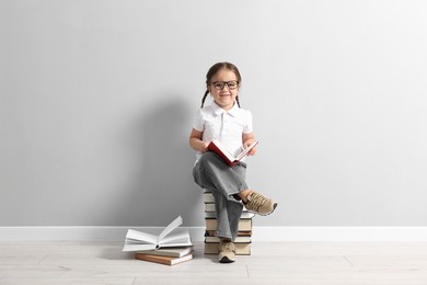 Photo of Cute little girl in glasses sitting on stack of books near light grey wall