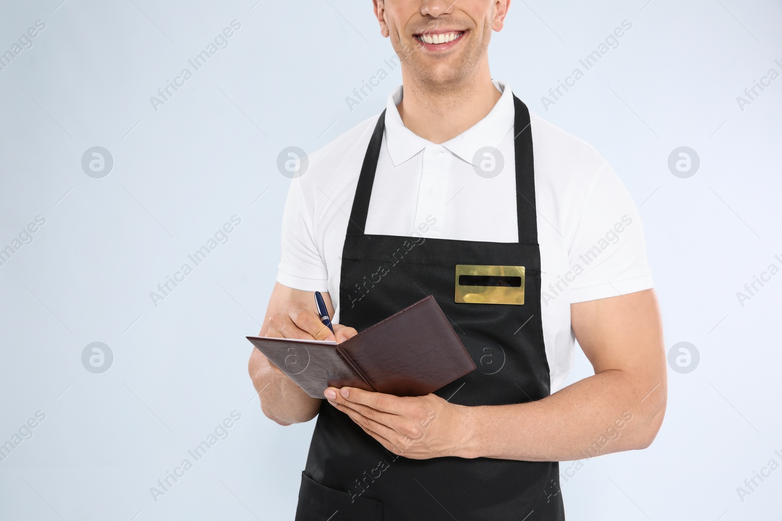 Photo of Young waiter in apron taking order on light background