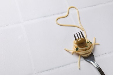 Photo of Heart made of tasty spaghetti, fork and olive on white tiled table. Space for text