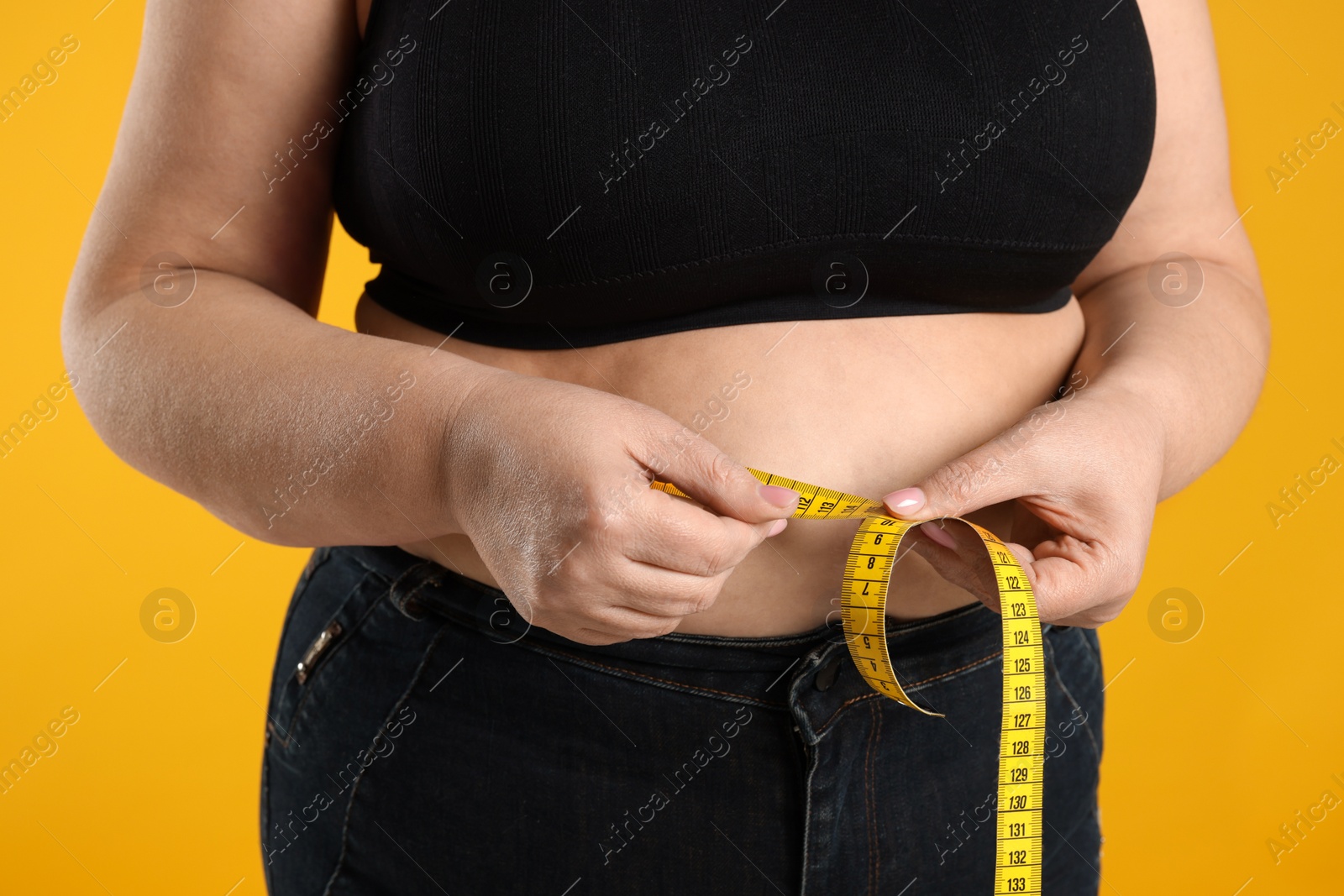 Photo of Overweight woman measuring waist with tape on orange background, closeup