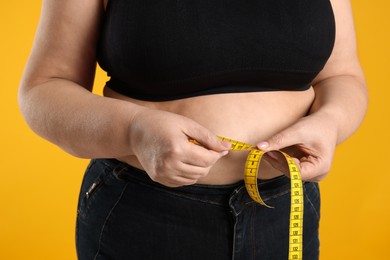 Photo of Overweight woman measuring waist with tape on orange background, closeup