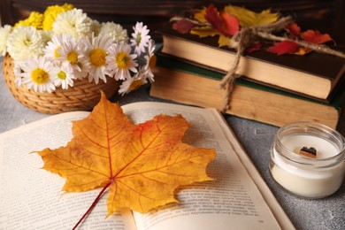 Photo of Book with autumn leaf as bookmark, scented candle and chamomile flowers on light gray textured table, closeup