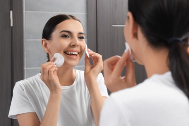Photo of Young woman using cotton pads with micellar water near mirror in bathroom