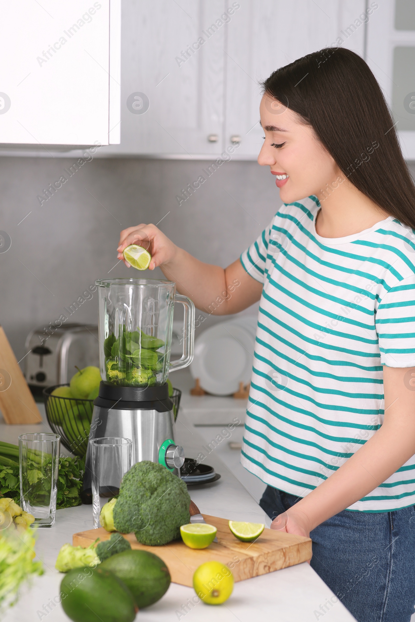 Photo of Beautiful young woman adding lime into blender with ingredients for smoothie in kitchen