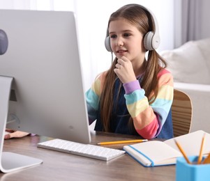 Photo of E-learning. Cute girl using computer and headphones during online lesson at table indoors