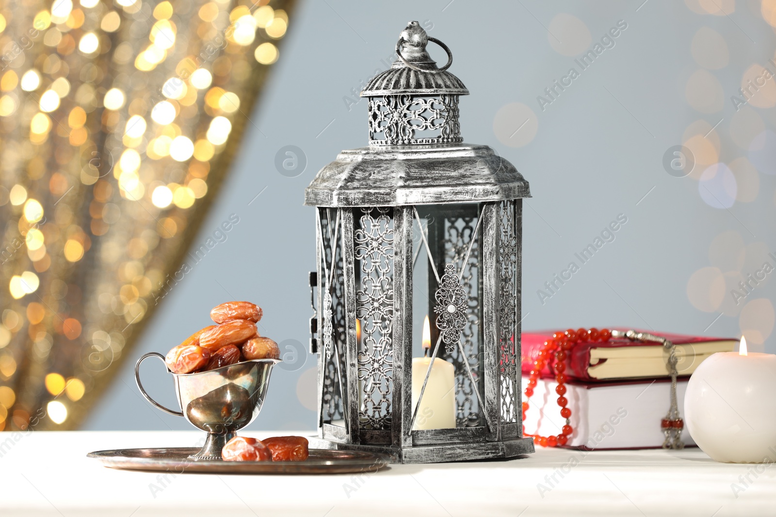 Photo of Arabic lantern, Quran, misbaha, candle and dates on white table against blurred lights