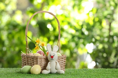 Photo of Cute Easter bunny toy with wicker basket and dyed eggs on green grass against blurred background, space for text