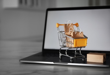 Internet shopping. Modern laptop and small cart with bags and box on marble table indoors, space for text