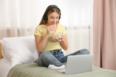 Photo of Teenage girl with headphones eating popcorn while using laptop on bed at home