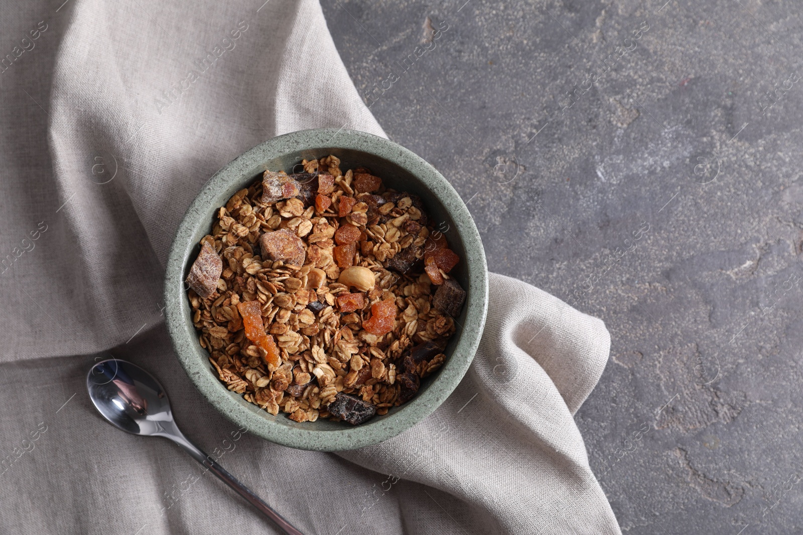 Photo of Tasty granola in bowl, spoon and napkin on gray textured table, flat lay. Space for text