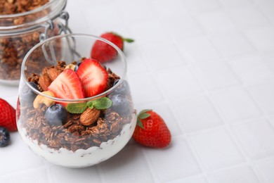 Photo of Tasty granola with berries, nuts and yogurt in glass on white tiled table, closeup. Space for text