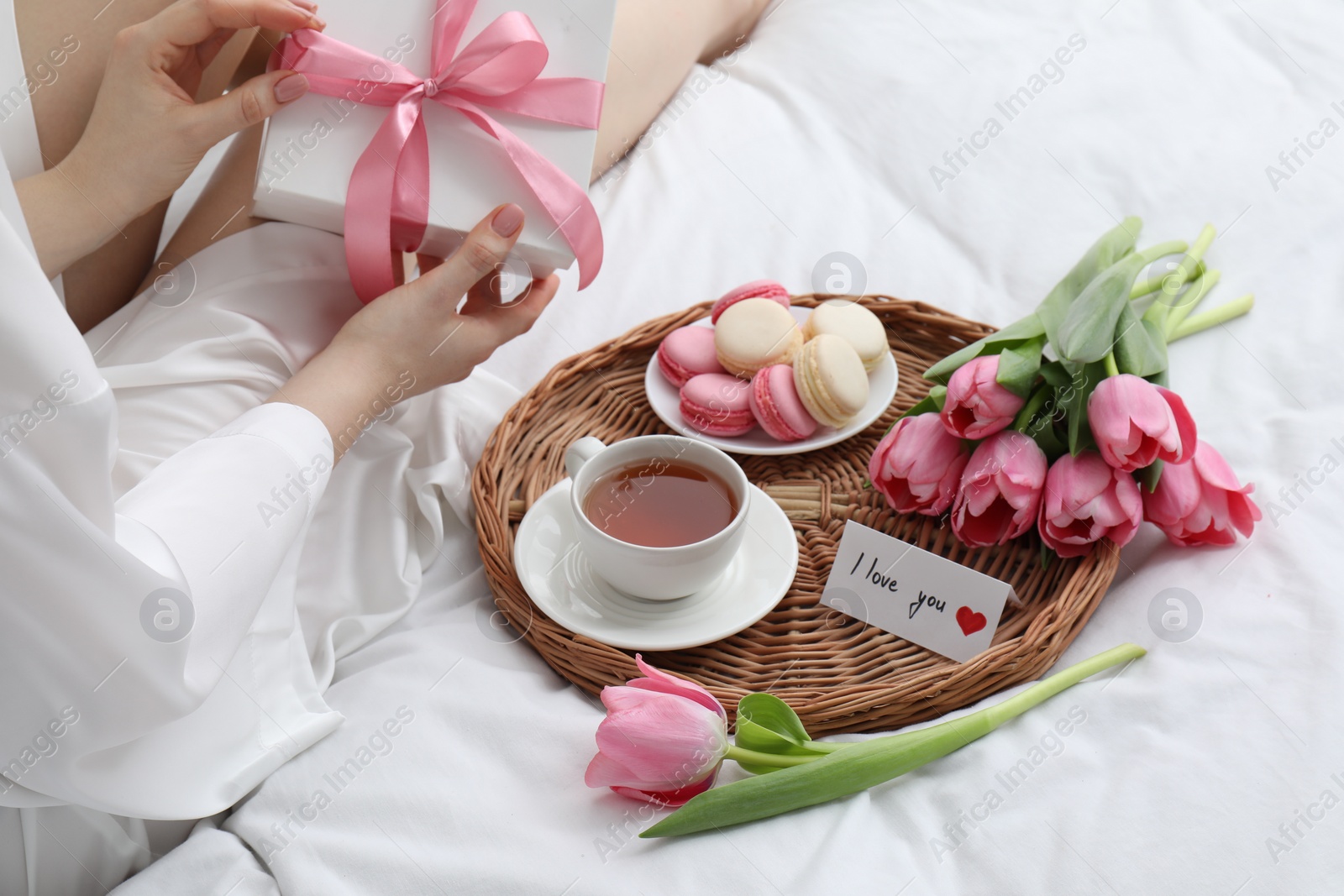 Photo of Tasty breakfast served in bed. Woman with gift box, tea, macarons, flowers and I Love You card at home, closeup