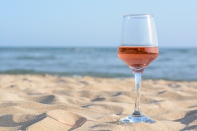 Photo of Glass of tasty rose wine on sand near sea, space for text