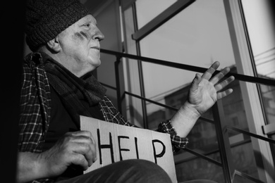 Photo of Poor senior man with cardboard sign HELP on stairs indoors. Black and white effect