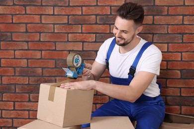 Photo of Smiling worker taping box with adhesive tape dispenser near brick wall