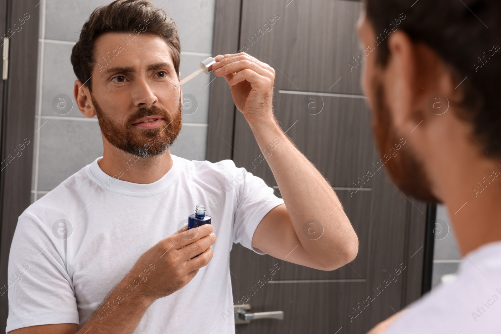 Photo of Handsome man applying oil on beard in bathroom