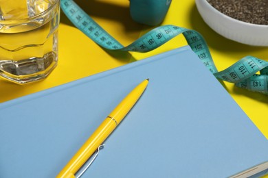 Photo of Weight loss concept. Composition with notebook, measuring tape and glass of water on yellow background, closeup