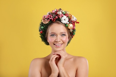 Beautiful young woman wearing Christmas wreath on yellow background