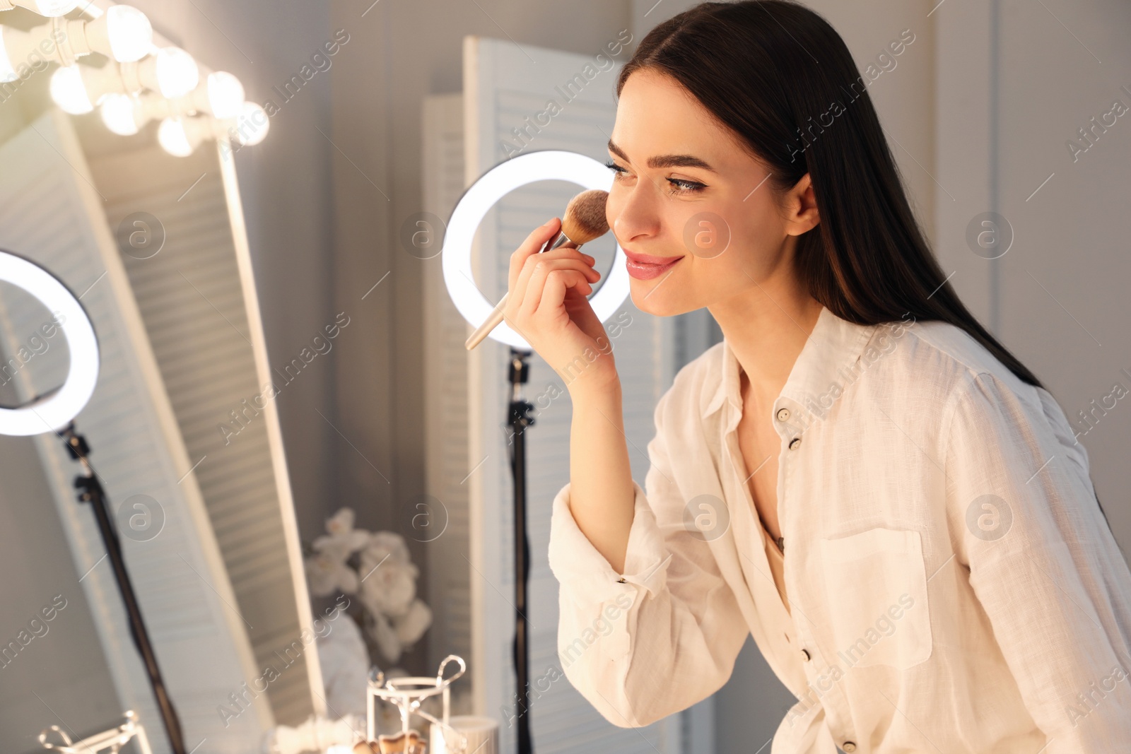 Photo of Young woman applying make up near illuminated mirror indoors