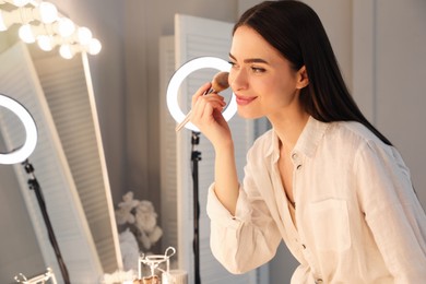 Young woman applying make up near illuminated mirror indoors