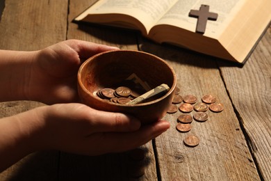 Photo of Donate and give concept. Woman holding bowl with coins and dollar banknotes at wooden table, closeup