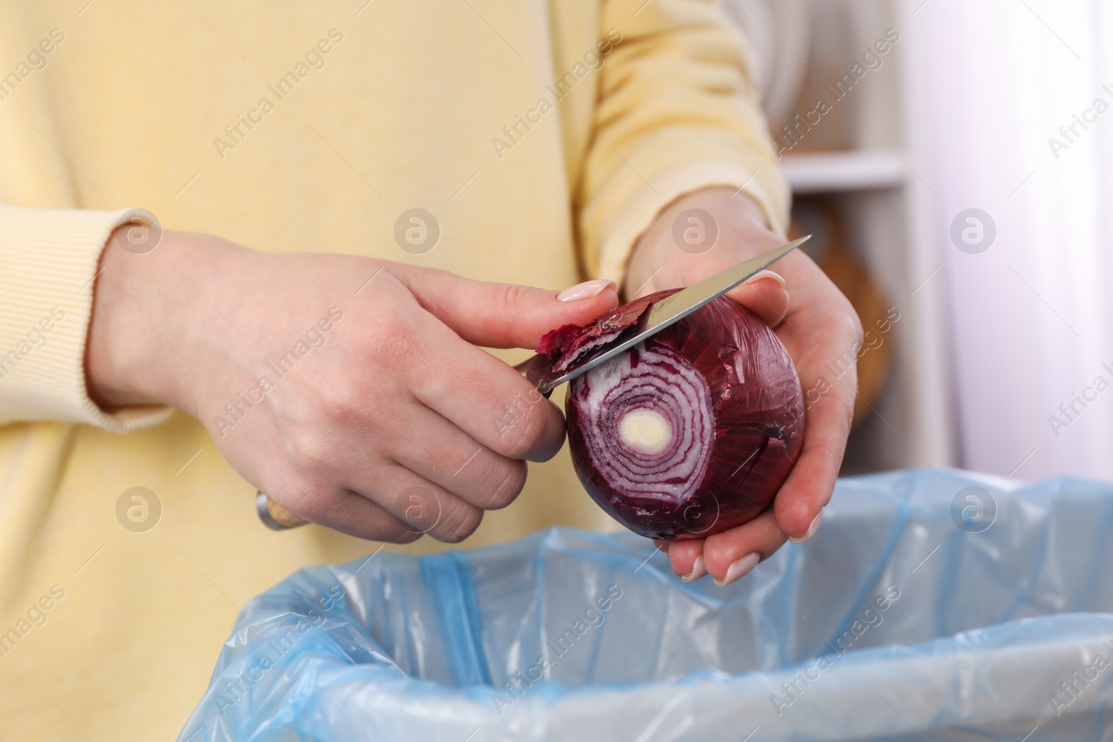 Photo of Woman peeling fresh onion with knife above garbage bin indoors, closeup