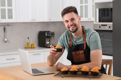 Photo of Happy man holding muffin near laptop at table in kitchen. Time for hobby