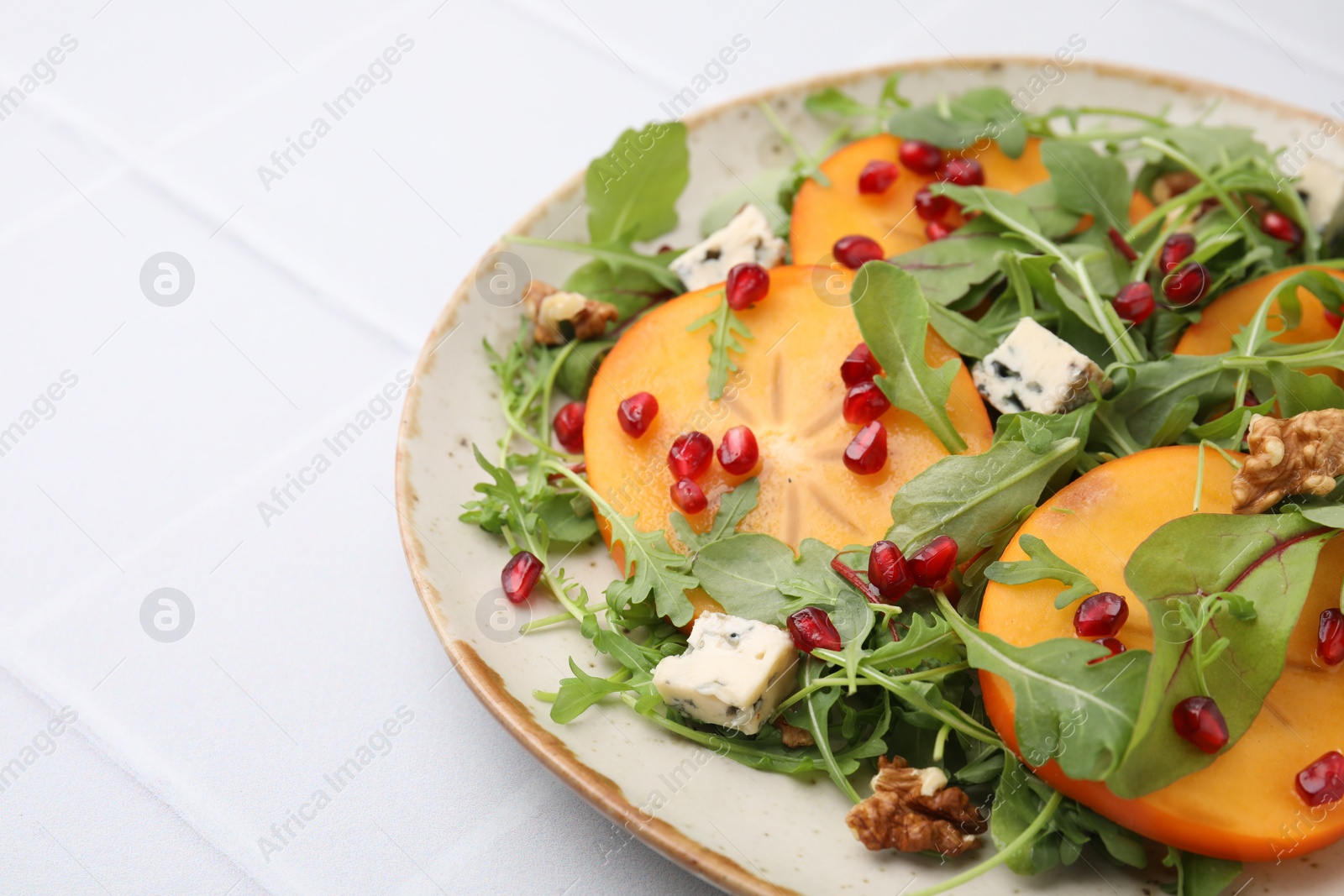 Photo of Tasty salad with persimmon, blue cheese, pomegranate and walnuts served on white tiled table, closeup. Space for text