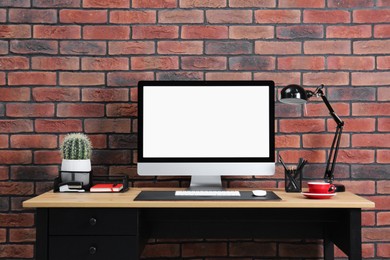 Photo of Stylish workplace with computer, lamp and stationery on wooden table near brick wall