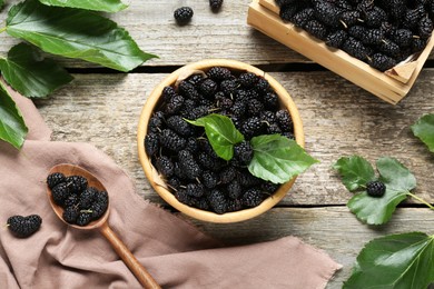 Bowl, spoon and crate of delicious ripe black mulberries on wooden table, flat lay