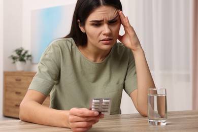 Photo of Sad woman with blister of pills suffering from headache at wooden table indoors