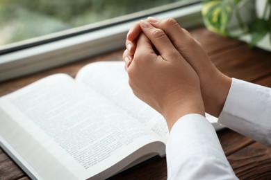 Photo of Woman holding hands clasped while praying at wooden table with Bible, closeup. Space for text
