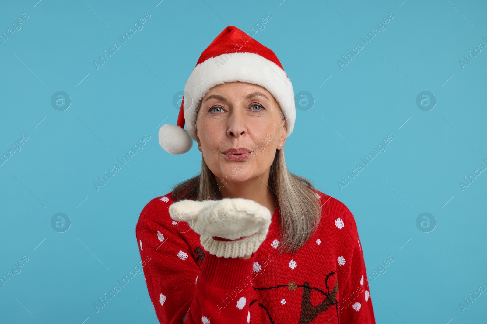 Photo of Senior woman in Christmas sweater and Santa hat blowing kiss on light blue background