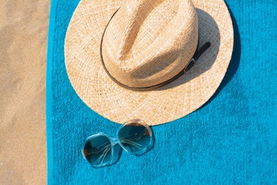 Soft blue beach towel with straw hat and sunglasses on sand, flat lay