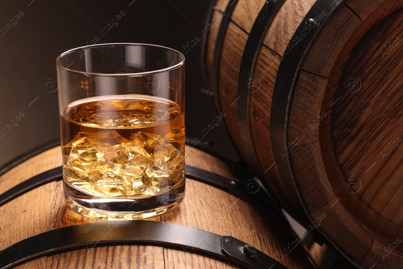 Photo of Whiskey with ice cubes in glass on wooden barrel against dark background, closeup