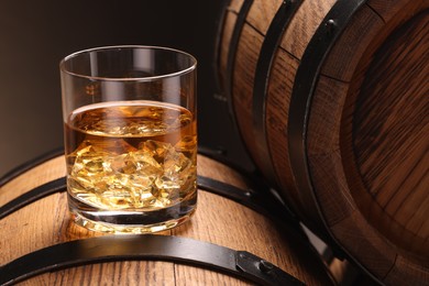 Whiskey with ice cubes in glass on wooden barrel against dark background, closeup
