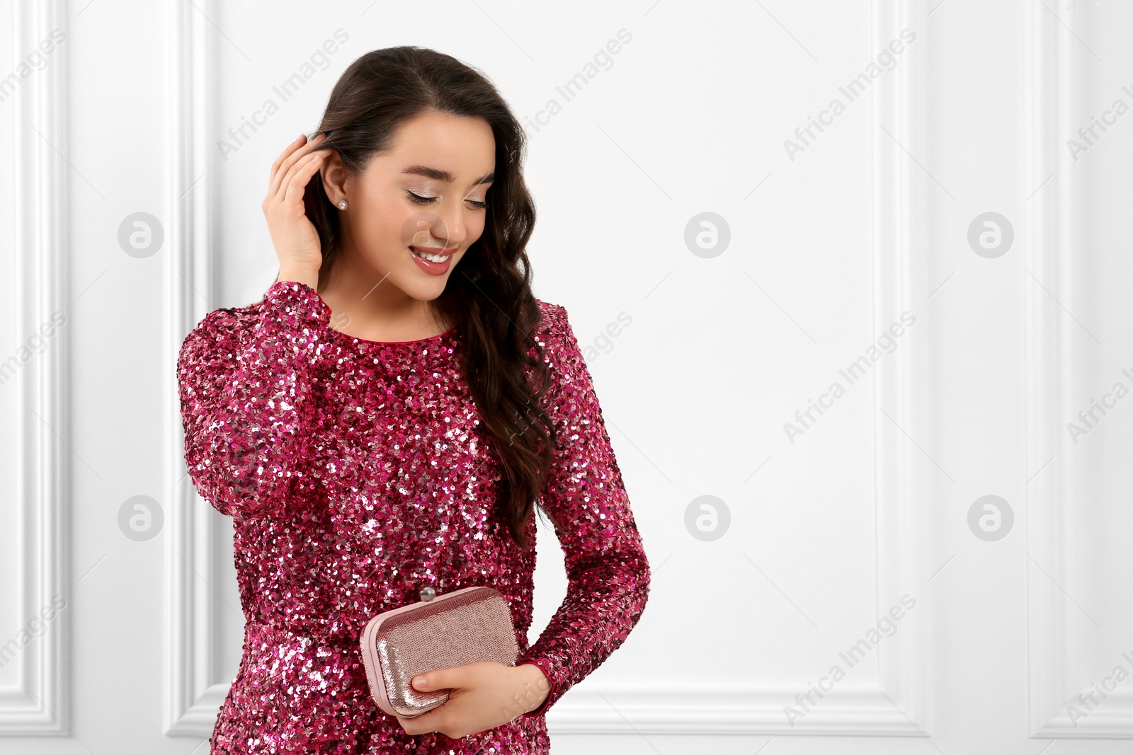 Photo of Beautiful young woman with clutch in stylish pink sequin dress near white wall indoors, space for text. Party outfit