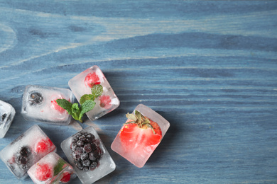 Photo of Ice cubes with different berries and mint on blue wooden table, flat lay. Space for text